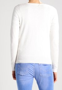 Valkoinen neulottu villapaita, jossa on pieniä pisteitä muodostava teksturoitu kuvio, pyöreä pääntie ja pitkät hihat, yhdistetty vaaleansinisiin farkkuihin.