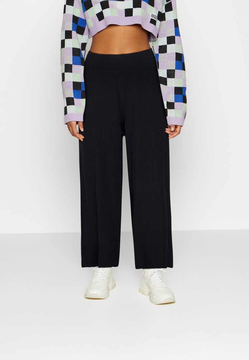 Femme portant un pantalon noir à jambes larges, des baskets blanches et un pull court à manches longues à carreaux aux couleurs noire, bleue, lavande et menthe.