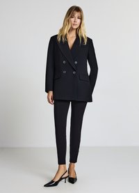 Schwarze, doppelt geknöpfte Blazer mit großen Knöpfen, kombiniert mit schmalen schwarzen Hosen und spitzen Slingback-Absätzen, alles in einem taillierten Design.