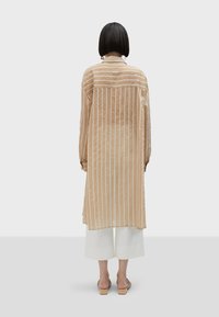 Ljusbeige lång skjorta med vertikala vita ränder, knappad framtill, något genomskinligt tyg och avslappnad passform, tillsammans med vita culotter och sandaler.