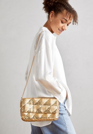 Vrouw draagt een witte blouse en blauwe spijkerbroek en heeft een gouden, met pailletten versierde, gewatteerde handtas met een kettingriem over haar schouder hangen.