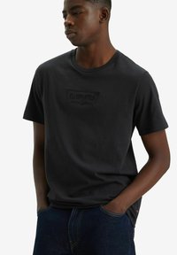 Camiseta de algodón negra con un logo texturizado de Levi's en el pecho, cuello redondo y mangas cortas, combinada con jeans de mezclilla azul.