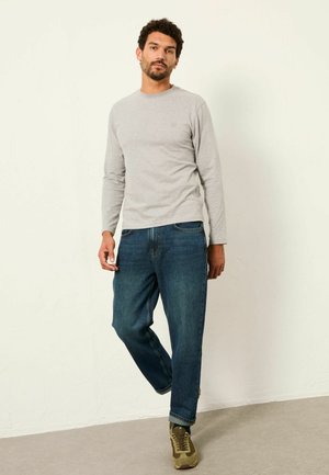 Mies seisoo beigeä seinää vasten, päällään vaaleanharmaa pitkähihaispaita, siniset farkut ja oliivinvihreät tennarit valkoisilla pohjilla.