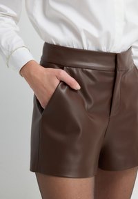 Mano in tasca dei pantaloni corti in pelle marrone a vita alta, indossati con una camicia bianca a maniche lunghe e collant neri trasparenti su uno sfondo chiaro.