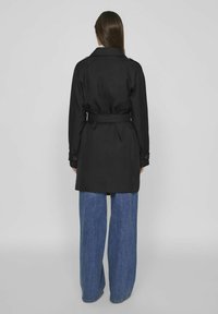 Juodas trenčkotis su juosmeniu diržu, ilgomis rankovėmis ir užsegamais rankogaliais, derinamas su plačiomis mėlynomis džinsomis. Lygus audinys, švarios linijos.