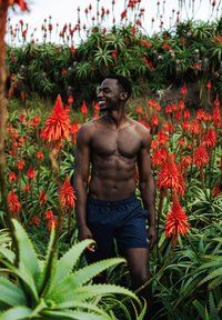 Een shirtloze man in blauwe shorts staat te midden van hoge rode bloemen en groene bladeren. Hij heeft een gespierd postuur, natuurlijk zonlicht en een groene achtergrond.