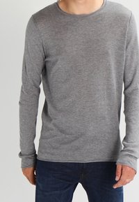 Homme portant un t-shirt gris uni à manches longues et col rond, avec un jean bleu foncé, debout devant un fond neutre.