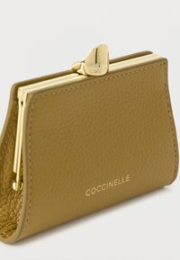 Portofel pentru monede din piele verde măsliniu, cu un clește de tone aurii, prezentând o textură netedă și logo-ul "Coccinelle" embosat pe partea din față.