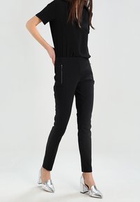 Pantalons ajustés noirs avec poche zippée, associés à un haut noir à manches courtes et des chaussures à talons carrés argentées à bout pointu.