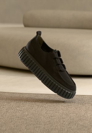 Zwarte casual sneaker met dikke geribde zool die boven een beige tapijt zweeft in een moderne kamer met neutrale tinten.