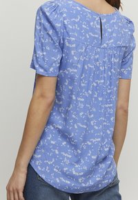 Blusa azzurra con motivo floreale, maniche a sbuffo corte, chiusura con bottone sul retro, e una vestibilità ampia con plissè sul retro.