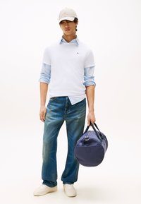 Bela majica s kratkimi rokavi preko svetlo modre srajce z ovratnikom, modre kavbojke, beli supergi in bež kapa; v roki drži temno modro torbo.