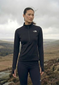 Frau steht an einem bewölkten Tag draußen und trägt eine schwarze The North Face Reißverschlussjacke und Leggings in einer rauen, hügeligen Landschaft.
