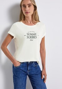 Vit bomullströja med rullade ärmar, med en grön grafisk tryck som s�äger "SUNDAY SCRIBES Club." Bärs med blå denimjeans.