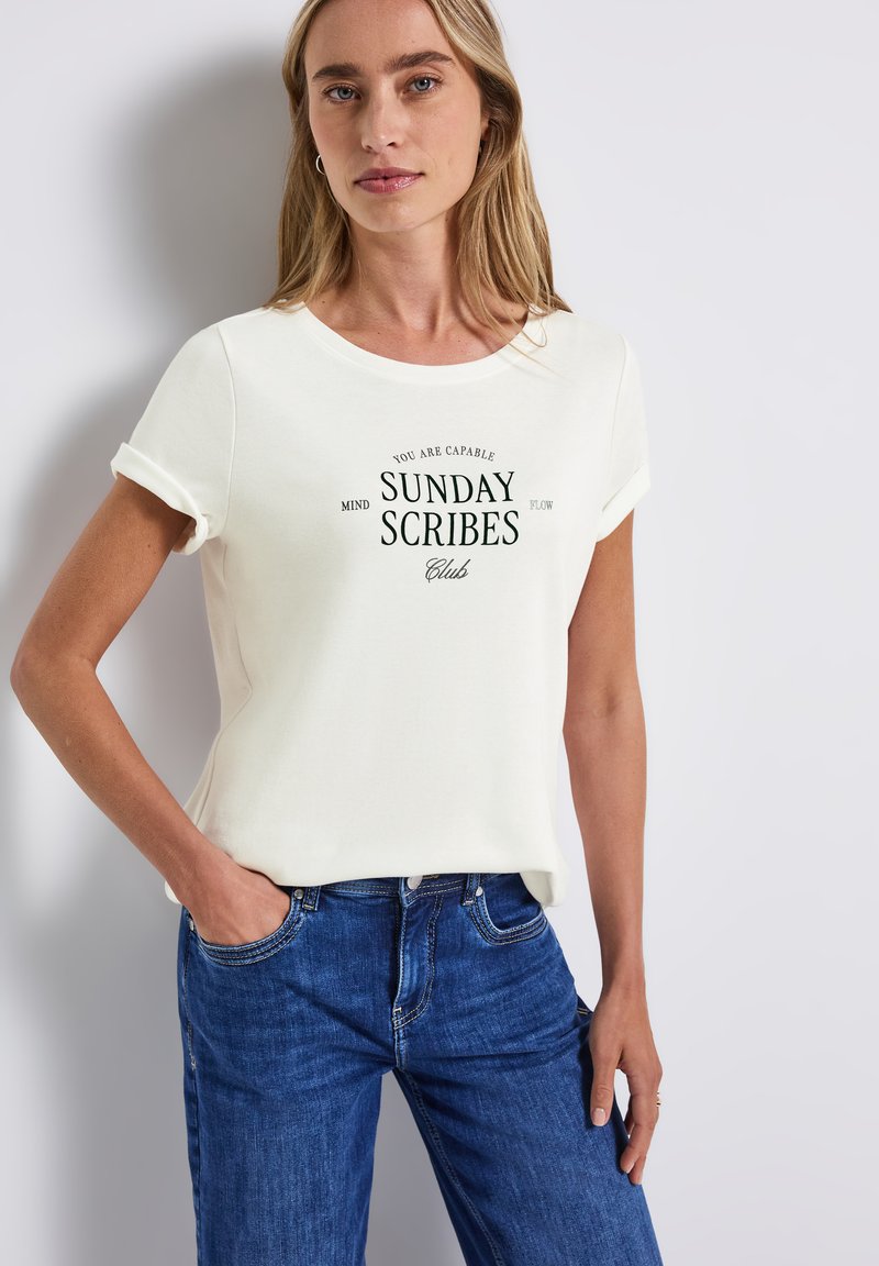 Vit bomullströja med rullade ärmar, med en grön grafisk tryck som säger "SUNDAY SCRIBES Club." Bärs med blå denimjeans.
