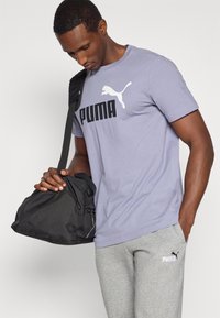 Λιλά μπλουζάκι με μεγάλο μαύρο τύπωμα "PUMA" και λογότυπο, σε συνδυασμό με γκρι φόρμα με μικρό λογότυπο. Μαύρη τσάντα duffel με χερούλια.