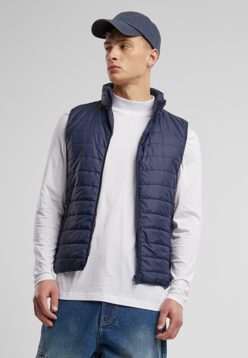 Marineblauer gesteppter Weste mit hohem Kragen, Frontreißverschluss, getragen über einem weißen Langarmshirt, kombiniert mit einer passenden marineblauen Mütze. Lässiges Design.