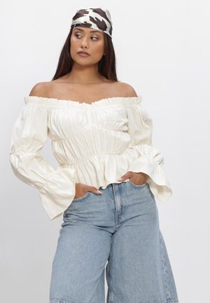 Vrouw draagt een witte off-shoulder blouse, lichtblauwe jeans met hoge taille en een zwart-witte patroon hoofdband, staand met handen in de zakken.