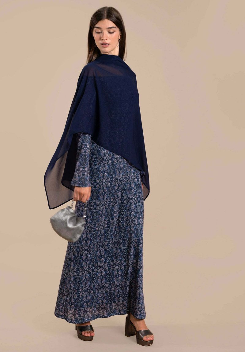 Vestido maxi azul marino con estampado floral y un sobrecapa transparente, que presenta un diseño estampado, combinado con un pequeño bolso plateado y tacones negros gruesos.