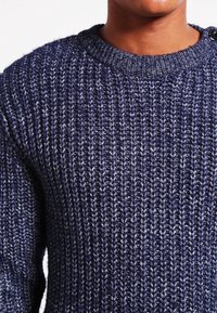 Marinblå stickad tröja med en texturerad mönstring, med rund halsringning och en knappdetalj på axeln.