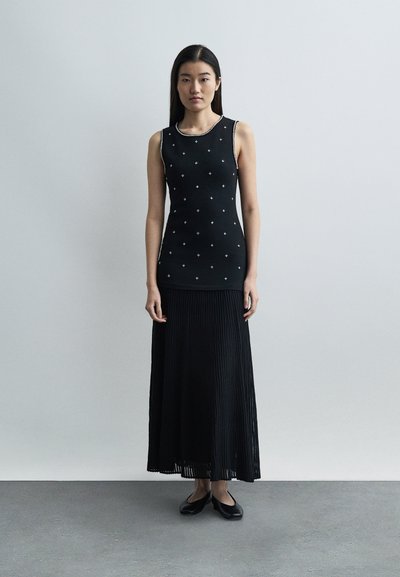 Νεαρή γυναίκα όρθια, φορεί μαύρο αμάνικο τοπ με λευκά σχέδια σταυρού, μακριά μαύρη πλισέ φούστα και μαύρες μπαλαρίνες, ουδέτερο φόντο.