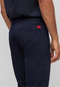 Navy blue cotton trousers with a slim fit, featuring a red logo tag on the back pocket and a smooth texture.