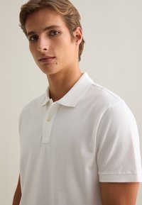 Camisa polo blanca hecha de algodón texturizado, con un cuello clásico, tapeta de dos botones y mangas cortas. Diseño simple y limpio.
