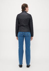 Svart quiltet jakke med høy krage, lange ermer og tettsittende silhuett, kombinert med blå rette jeans og svarte sko.