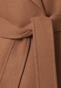Brązowy wełniany płaszcz o miękkiej fakturze. Posiada pasek z przodu do wiązania oraz kątowe klapy. Widok z bliska podkreśla materiał i szwy tkaniny.