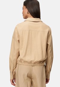 Lichtbeige cropped jack met een kraaghals, elastische tailleband en lange mouwen. Soepele stof met een relaxte pasvorm en knopenmanchetten.