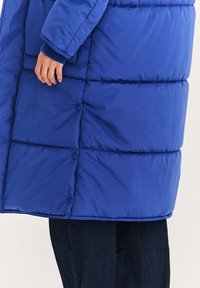 Niebieska pikowana kurtka puchowa z przeszywanymi panelami, zatrzaskami i ściągaczami na rękawach. Tkanina jest gładka i wodoodporna.