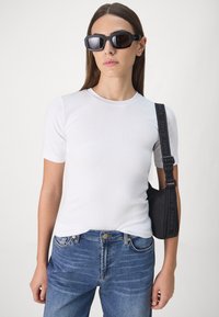 Hvit, tettsittende t-skjorte med korte ermer, blå denimjeans, svart crossover-veske med logo-rem, og svarte firkantede solbriller med mørke linser.
