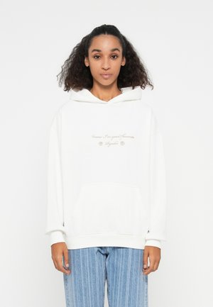 Hvit hettegenser med stor frontlomme, snorhette og lys beige tekst. Kombinert med blåstripete jeans, står modellen foran en ensfarget bakgrunn.