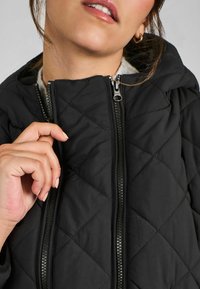 Veste matelassée noire avec un col montant, fermeture à double zip et une doublure intérieure douce. Couture détaillée visible sur la surface de la veste.