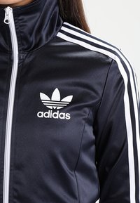 Chaqueta deportiva de satén azul marino con cuello alto, cremallera blanca y tres rayas blancas en cada hombro. Presenta un logo de Adidas en blanco.