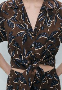 Brown short-sleeve shirt with a tie-front design, featuring blue and cream leaf patterns, and a collar with wooden button closures.