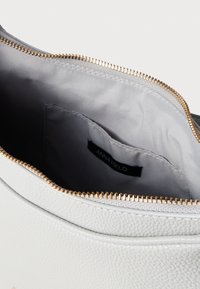 Fehér texturált kézitáska arany cipzárral. A belső rész szürke szövetből készült, és egy "ANNA FIELD" feliratú címkét tartalmaz. Sima, ívelt tetővel rendelkezik.