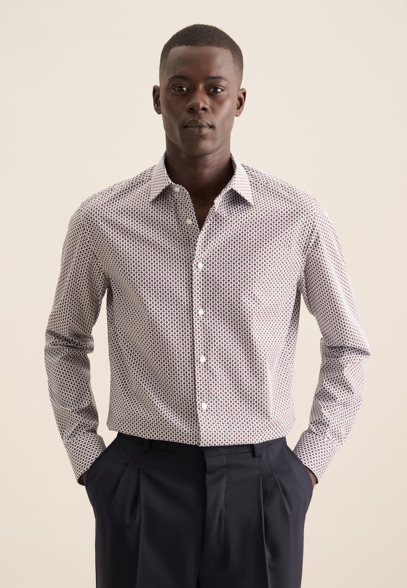 Camicia bianca con motivo geometrico rosso e blu navy, maniche lunghe e colletto classico. Abbinata a pantaloni blu navy scuri.