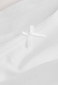 Tissu blanc avec une fine bordure en dentelle festonnée et un petit nœud en satin blanc centré, montrant des coutures détaillées et la texture.