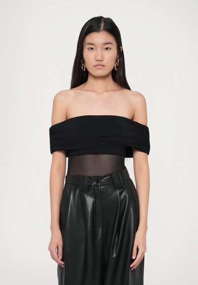 Giovane donna con lunghi capelli scuri che indossa un top nero scollato, con una sezione centrale trasparente, pantaloni in pelle neri a vita alta e grandi orecchini a cerchio d'oro.