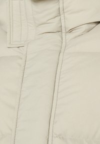 Beige pufferjacka med kviltad textur, med synlig sömnad, en hög krage och en framfickslåsning. Mjukt, lätt material.
