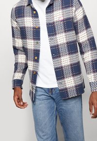 Camicia a scacchi blu e bianca con tessuto testurizzato, caratterizzata da cuciture a contrasto e bottoni marroni, indossata sopra una maglietta bianca e jeans.