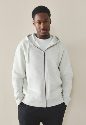 Young man standing against beige wall wearing light gray zip-up hoodie and black pants, hands in pockets, looking straight ahead.