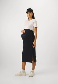 Trudnička odjeća koja uključuje usku crnu suknju sa bočnim prorezom, u kombinaciji s bijelom majicom kratkih rukava i bež tenisicama.