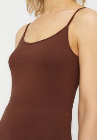 Κοντινό πλάνο ενός ατόμου που φοράει καφέ κορσέ με λεπτές τιράντες, δείχνοντας τον πάνω κορμό και τους ώμους.