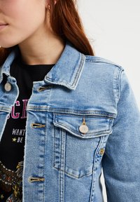 Ljusblå denimjacka med spetsig krage, bröstficka och silverdetaljer. Har gula sömmar och tyget är något slitet.