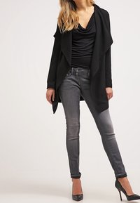 Černý otevřený kardigan s přehozeným límcem, kombinovaný s upnutými šedými džíny a černými podpatky. Hladká textura a moderní střih.