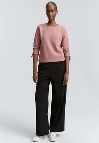 Rozā faktūras adīts džemperis ar trim ceturtdaļas piedurknēm un saistītiem manžetēm, kombinēts ar augsta vidukļa melnām platām biksēm un baltām sporta kurpēm.