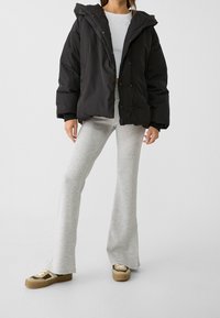 Schwarze Pufferjacke mit Kapuze, mit einem Knopfverschluss und Seitentaschen. Kombiniert mit hellgrauen, ausladenden Jogginghosen und beigen Sneakers.