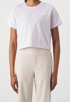 Persoon draagt een lichtpaarse cropped T-shirt met het logo van "The North Face" en beige broek met hoge taille tegen een effen achtergrond.